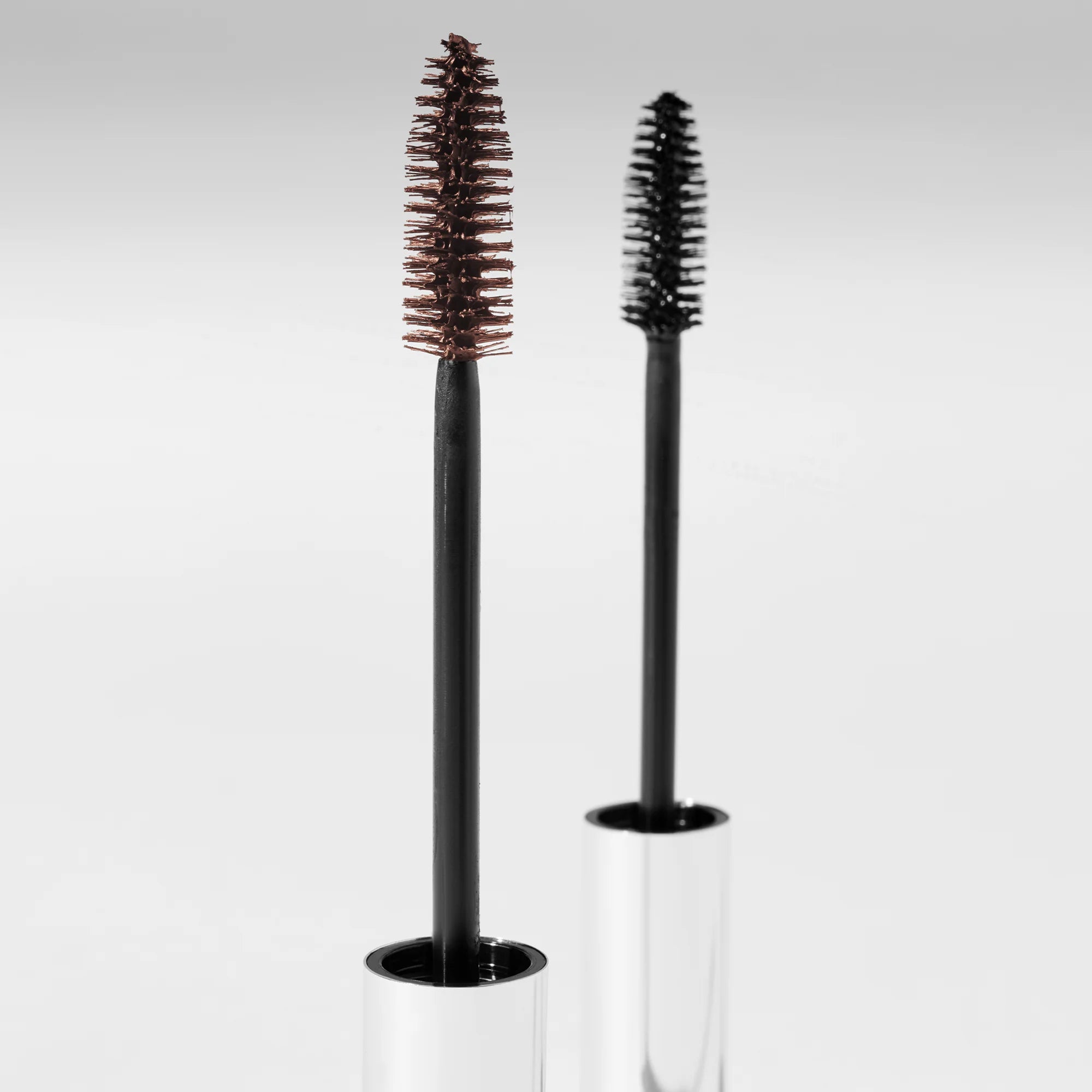 Two black mascara brushes with clear cylindrical holders on a light gray background
