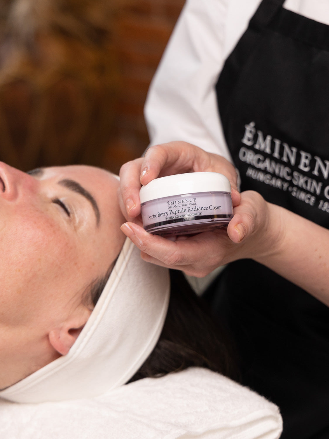 Person holding Eminence Organic Skin Care jar over another person's face