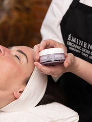 Person holding Eminence Organic Skin Care jar over another person's face