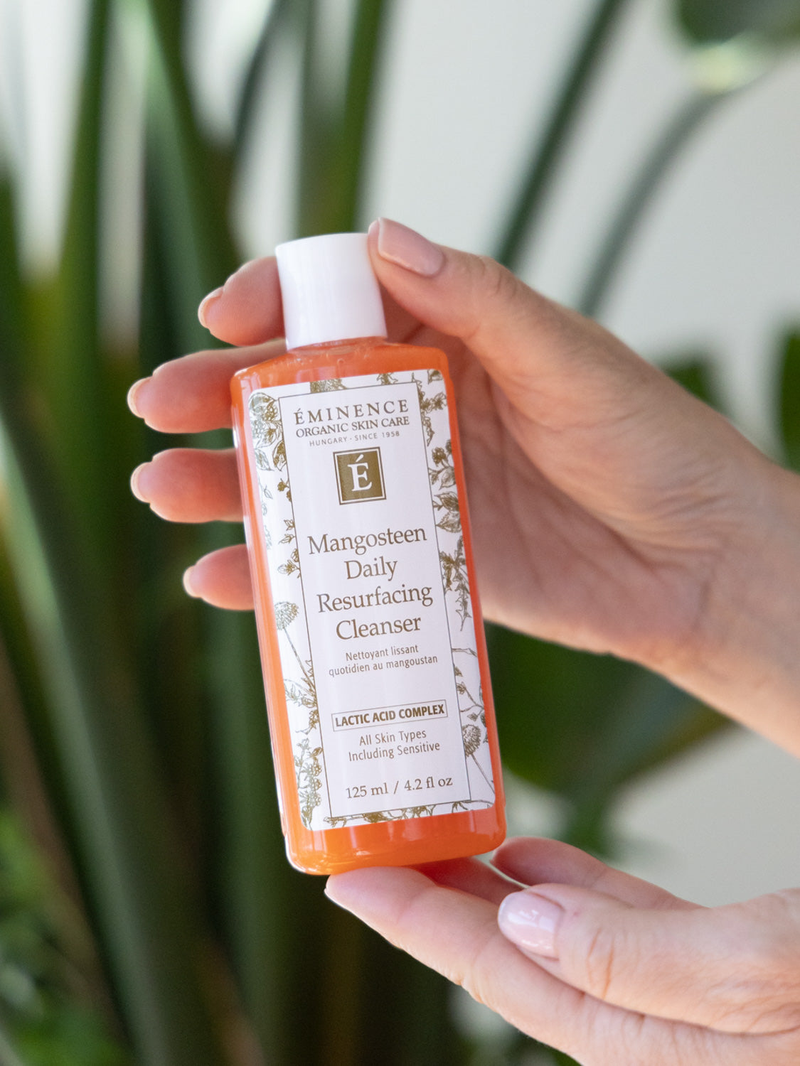 Hand holding a bottle of Eminence Organics Mangosteen Daily Resurfacing Cleanser with greenery in the background.