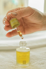 Hand using a dropper to apply facial recovery oil from a bottle labeled 'Facial Recovery Oil'.