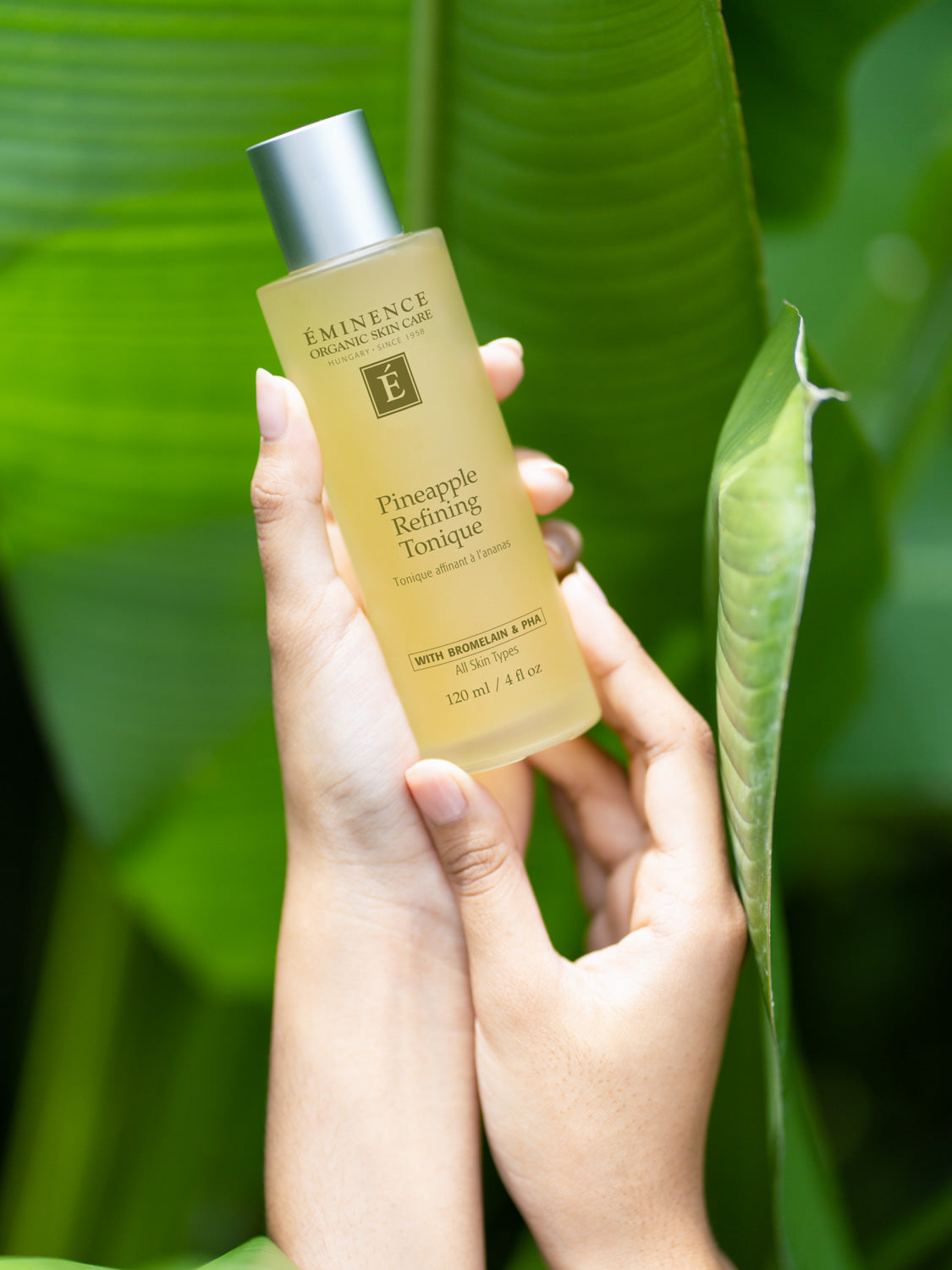 Hand holding a bottle of Eminence Pineapple Refining Toner against a green leafy background