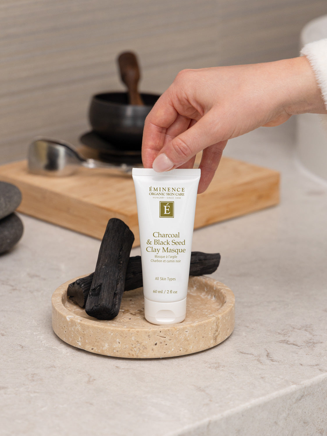 Tube of Eminence Charcoal & Black Seed Clay Masque on a wooden tray with charcoal pieces, held by a hand.