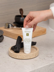 Tube of Eminence Charcoal & Black Seed Clay Masque on a wooden tray with charcoal pieces, held by a hand.