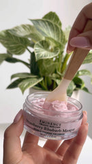 Hand holding a jar of Eminence Strawberry Khubarb Masque with a wooden applicator, plant in the background.