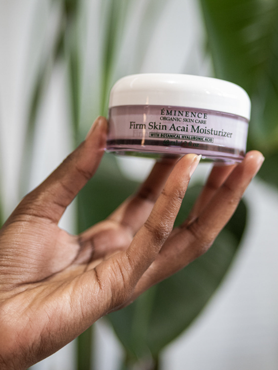 Hand holding a jar of Eminence Firm Skin Acai Moisturizer against a blurred green leafy background