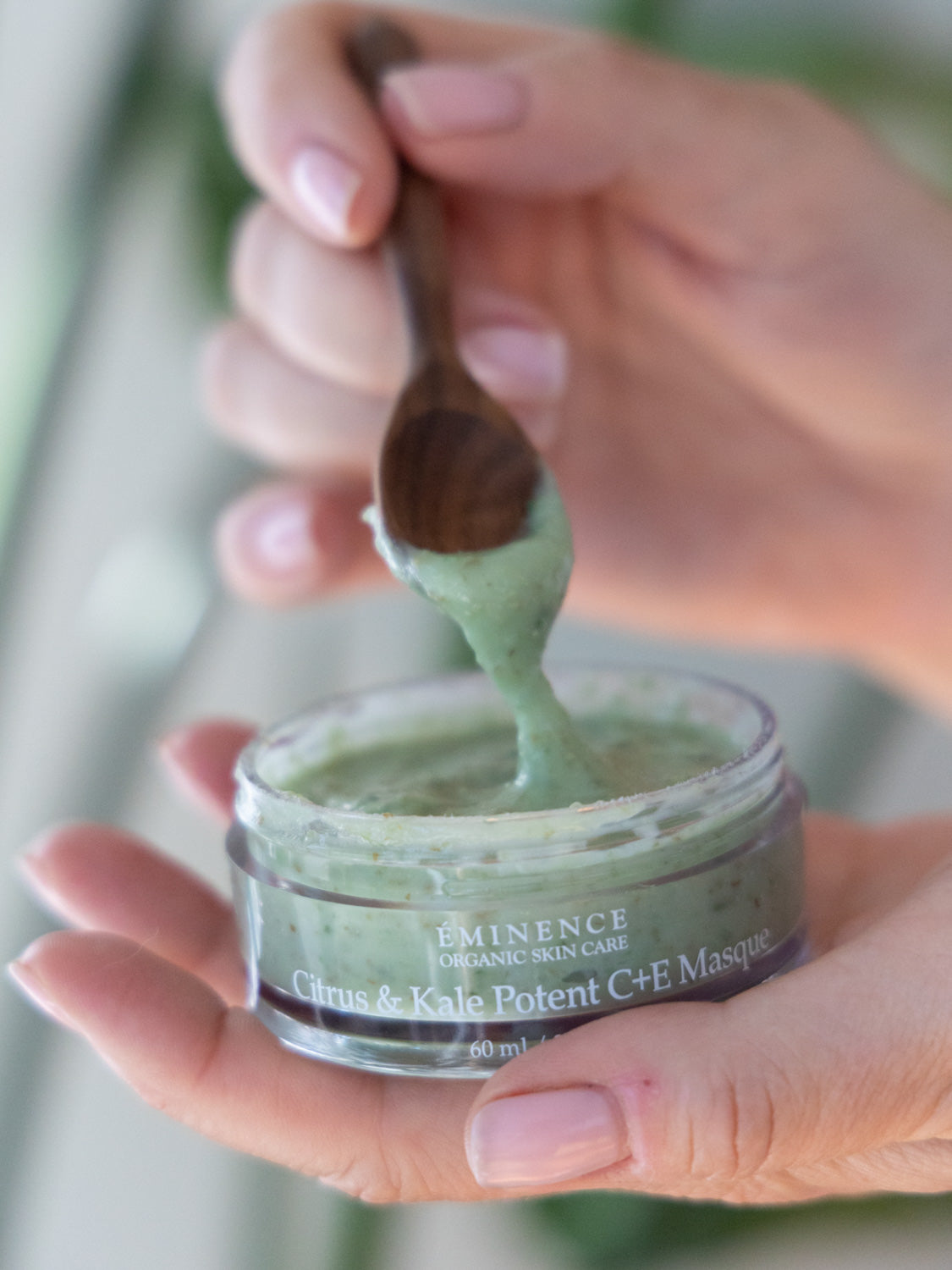 Hand holding a jar of Eminence Citrus & Kale Potent C+E Masque with a wooden spoon.