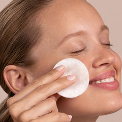 Woman applying a white cotton pad to her face against a neutral background